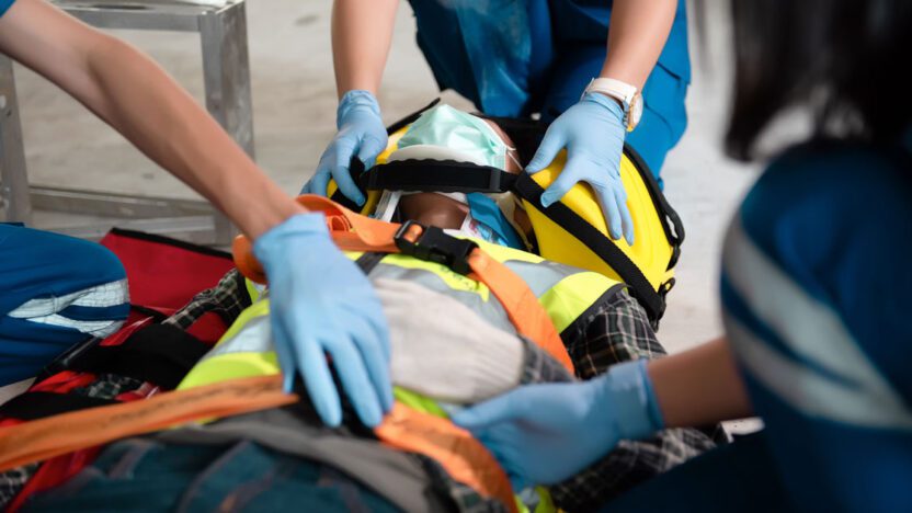 Medics in blue uniforms and gloves are attending to a person on a stretcher, securing them with straps and wearing safety gear, including a neck brace and a helmet.