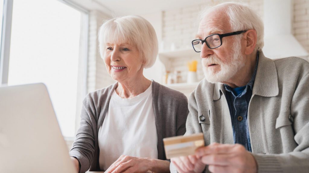 An elderly couple sits at a table, using a laptop. The man holds a credit card
