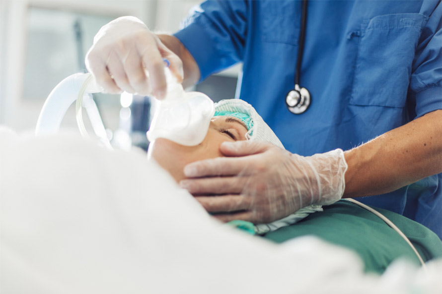 A medical professional in blue scrubs holds an oxygen mask to a patient's face in a clinical setting.