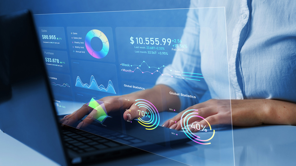 Person working on a laptop with digital financial dashboards displaying graphs, statistics, and data visualizations projected on a transparent screen.